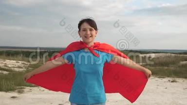 美丽的女超级英雄穿着红色斗篷在蓝天前穿过田野，微笑着慢动作。少女梦